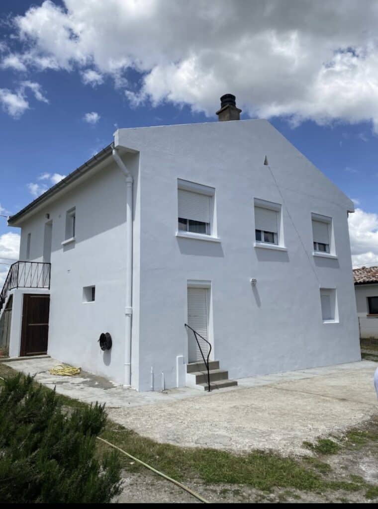 Maison blanche fraîchement peinte avec volets fermés, sous un ciel bleu nuageux. Escalier extérieur, porte de garage en bois, et terrain verdoyant.