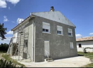 Maison en rénovation avec échafaudage. Façade grise inachevée, pignon blanc. Fenêtres avec volets roulants.
