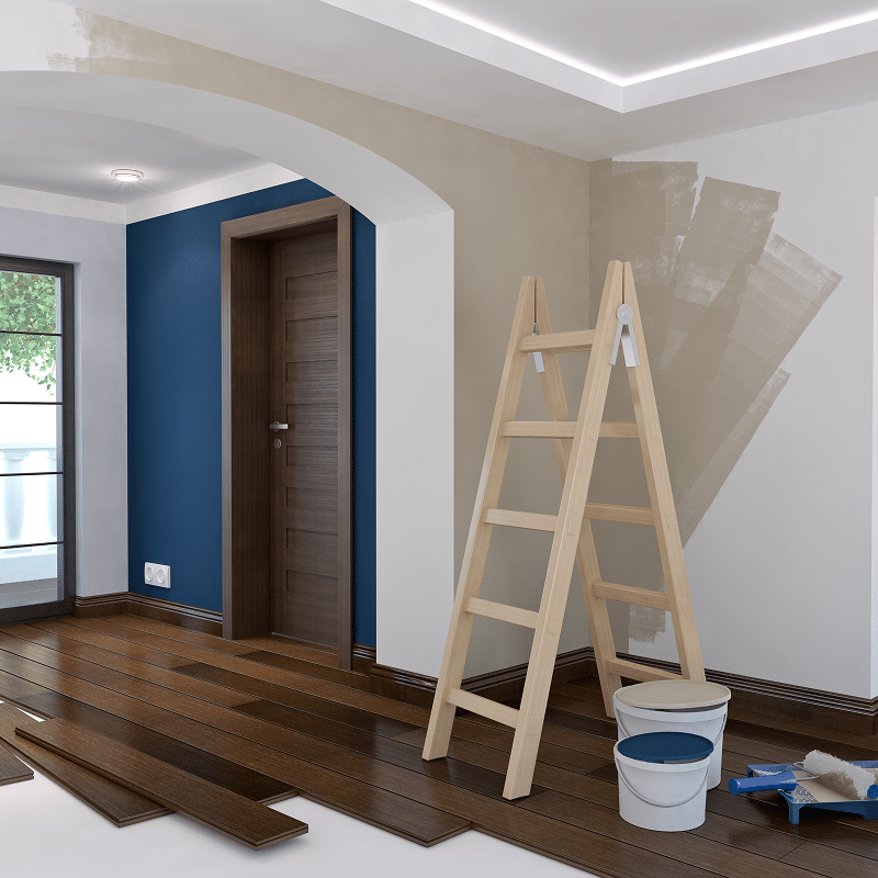 Intérieur en rénovation avec des murs en cours de peinture beige, un sol en parquet sombre en installation, et une échelle en bois. Deux pots de peinture et un bac à rouleau.