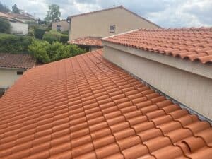 Toit de maison résidentielle en tuiles de terre cuite, orange-rouge et humides, avec en arrière-plan d'autres toits et un ciel nuageux.