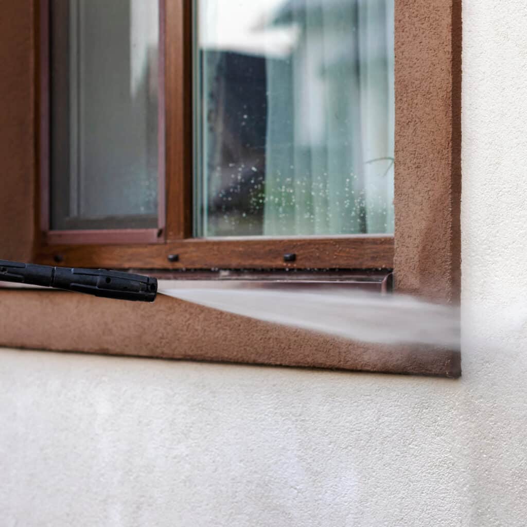 Nettoyage haute pression d'une façade de maison. Le jet d'eau atteint un mur beige texturé et un rebord de fenêtre marron sous une fenêtre.
