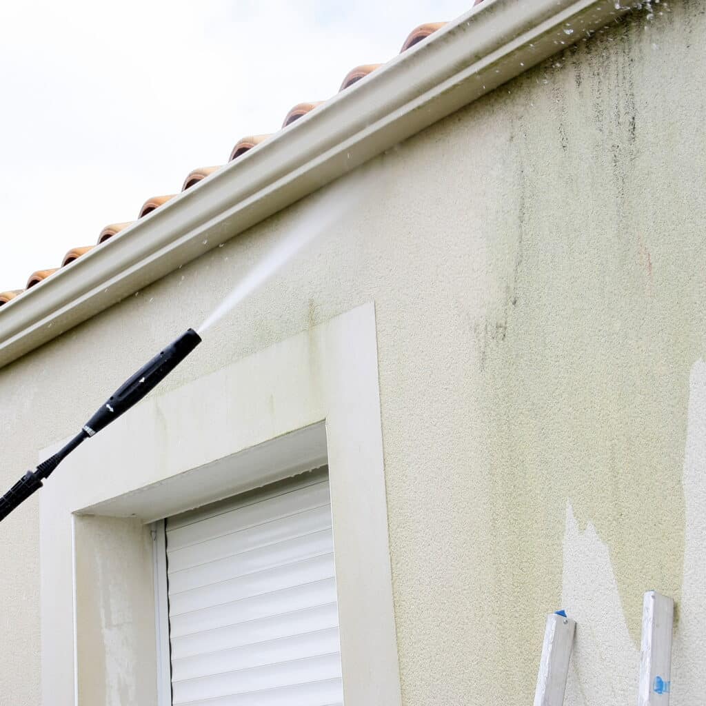 Nettoyage d'une façade de maison couverte de mousse avec un nettoyeur haute pression, révélant un mur propre.