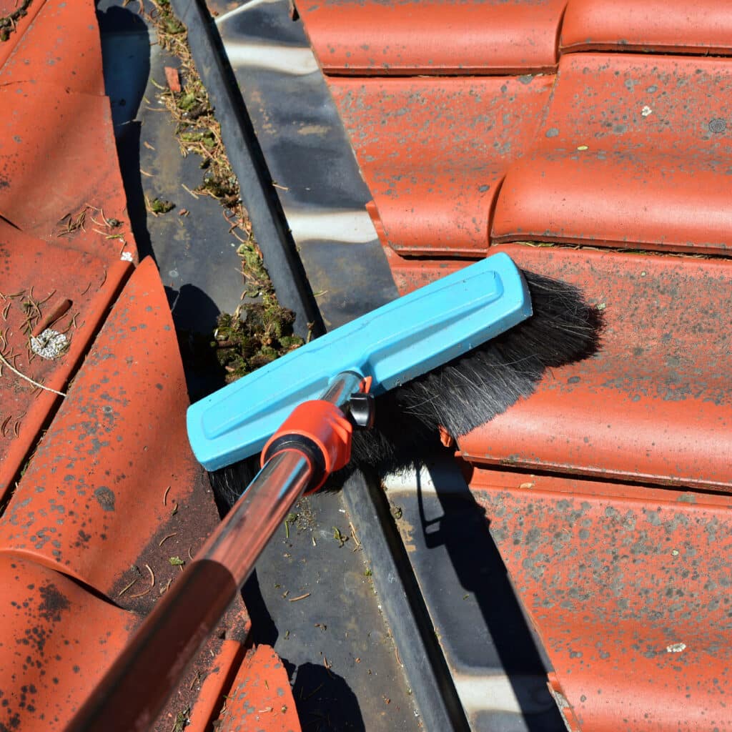 Une brosse bleue sur un long manche nettoie une gouttière noire remplie de débris (feuilles, mousse) le long de tuiles de toit rouges.