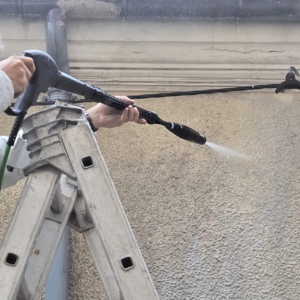 Mains nettoient un mur extérieur texturé avec un nettoyeur haute pression. Un jet d'eau puissant et une échelle en aluminium sont visibles.