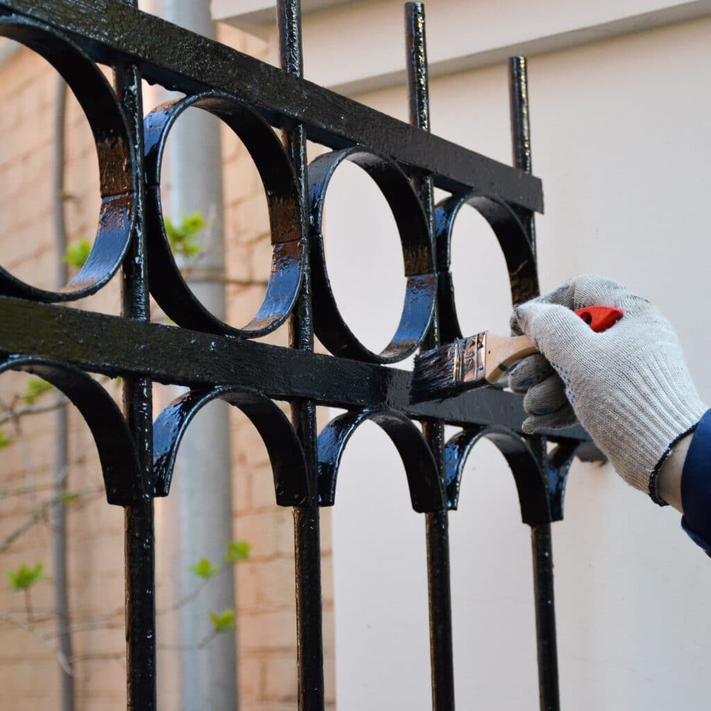 Main gantée peignant une clôture décorative en fer forgé noire avec un pinceau. La peinture noire est fraîche et brillante.