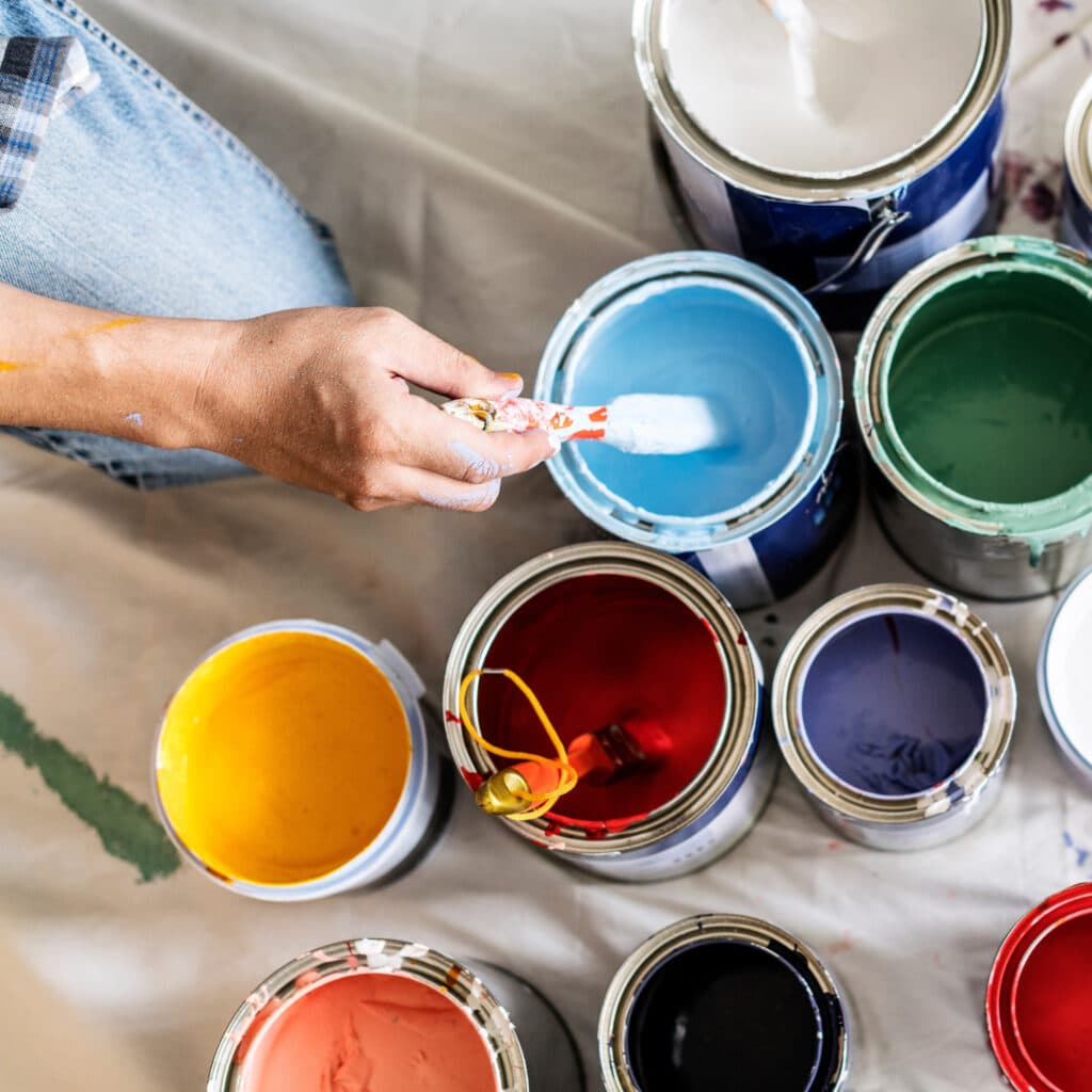 Une main trempe un pinceau dans un pot de peinture bleue. Plusieurs pots de couleurs vives (blanc, vert, jaune, rouge, violet) entourent la personne.